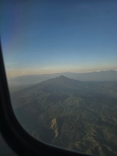 An aerial view captured through an airplane window shows a vast, verdant mountainous landscape under a clear blue sky. The foreground is dominated by the dark curve of the airplane window frame. The mountains, covered in dense forestation, stretch into the distance, becoming hazier and lighter in hue as they recede. In the middle ground, a prominent mountain peak stands out, its slopes also heavily forested. A distinct, lighter-colored scar or path is visible on one of the mountain slopes. The horizon is marked by a series of layered mountains and a faint haze that softens the distant view. The scene is devoid of any visible human presence or activity. The image does not provide any specific details that would allow for identification of the location as Ujongmesjid, Indonesia.