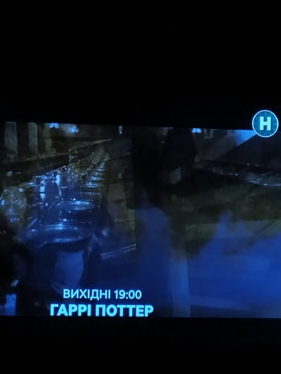 The image displays a screen showing a preview of a movie. In the upper right corner, a circular logo with the letter "H" is visible. Below this, text reads "ВИХІДНІ 19:00" which translates to "WEEKEND 19:00". Further down, the title "ГАРРІ ПОТТЕР" is displayed in large, bright blue letters, indicating "HARRY POTTER". The background of the screen shows a scene that appears to be from the movie, featuring multiple large pots or cauldrons in what looks like a dimly lit, outdoor environment. There are no discernible people in the foreground, and the overall scene is dark with blue and green hues. The image itself is a photograph of a screen, with a black border around it. There is no information within the image to suggest its location in Turka, Ukraine.