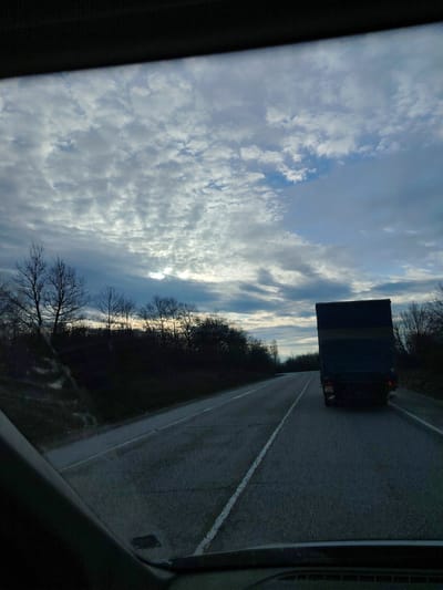 The image is a view from inside a vehicle, looking out the front windshield onto a rural road. The road has a white line dividing the lanes. A large truck is directly in front of the vehicle in the right lane. The roadside consists of trees and low vegetation. The sky is overcast with scattered clouds. The scene takes place near Iastrebino, Bulgaria.