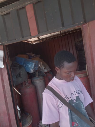 A man with short black hair lies on his side in a dark, cluttered space with industrial gas tanks of varying sizes and colors, bags, and other indistinguishable objects. The man wears a light pink t-shirt with a large design and a shoulder bag strap. The space is enclosed by metal structures, with the metal on the left side appearing to be painted in alternating dark gray and red stripes. Given the context of Eka, Nigeria, as provided in the prompt, the objects contained in the space might be related to gas distribution or industrial activities.