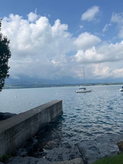 The image captures a daytime scene on Lake Thun, viewed from the shoreline in Hünibach, Switzerland. The sky features a mix of bright blue patches with white cumulus clouds, while darker, more uniform clouds obscure the upper portions of the distant mountains. The water surface displays moderate ripples, indicating slight movement.

In the immediate foreground, a dark grey concrete retaining wall or low pier extends horizontally into the water from the lower-left. The shoreline beneath this structure and to its right consists of dark grey rocks and sparse green vegetation. A dark green tree or bush partially frames the scene on the far left.

On the water, a single white sailboat with a prominent mast is anchored in the mid-right portion of the frame; its sails are furled. A second, partially visible white boat is located on the far right edge. Two white, spherical buoys are present in the water: one centrally located on the left side, and a smaller one further right. A single white swan is observed swimming near the left-most buoy. The background reveals a continuous line of green hills and mountains, with discernible residential structures scattered along the distant lakefront. No human subjects are present in the scene.
