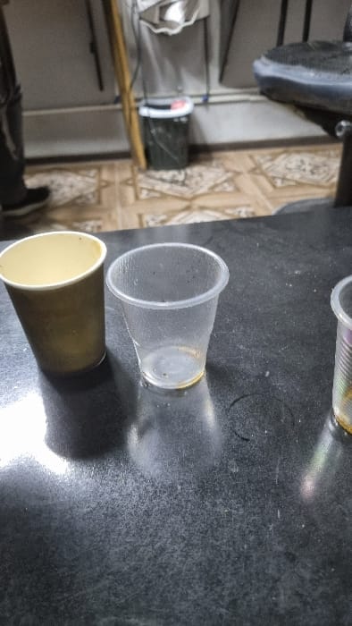 The scene depicts an interior setting in the city of Yangi-Khayat, Uzbekistan. In the foreground, a dark, textured, and somewhat reflective surface, likely a table or counter, holds three disposable cups. From left to right, these include a light-brown paper cup, an empty clear plastic cup with yellowish residue at its base and small dark specks inside, and a partially visible, ribbed clear plastic cup also containing yellowish residue. The background reveals a light-colored tiled floor with a repeating geometric pattern, meeting a light-colored wall. A wooden pole leans vertically against the wall, adjacent to a dark, cylindrical trash can with a white rim and a red detail. Above the trash can, multiple electrical wires are visible. On the far left, the lower calf and foot of an individual, wearing dark pants and a dark shoe with a white sole, are present on the tiled floor. A partial black, textured object, possibly part of a chair, is visible on the far right of the background. No direct actions or interactions are occurring.