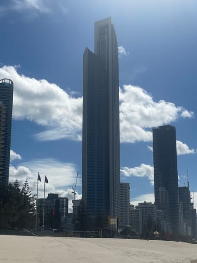 The image captures a view of the Surfers Paradise skyline in Australia, facing inland from a sandy beach under a partly cloudy sky. Dominating the center is the tall, slender skyscraper "Peppers" with its name visible at the top. To its left, another skyscraper with a rounded facade and a balcony structure is partially visible. To the right of Peppers, a dark, rectangular skyscraper stands, and further to the right, a series of other high-rise buildings form a dense urban backdrop. Several flags are planted on poles on the left side of the image, and a construction crane is visible among the buildings, suggesting ongoing development. In the foreground, the sandy beach is empty except for a few scattered individuals and beach infrastructure like a volleyball net. The scene depicts a bright, sunny day with scattered white clouds against a blue sky.