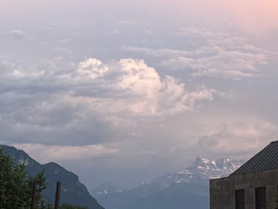 The image depicts a panoramic view of the mountains and clouds from Montreux, Switzerland. In the foreground, a building with a dark, angled roof and a stone facade is visible on the right side. The building has dark, slatted windows. To the left, lush green foliage from trees can be seen, along with two vertical wooden posts. In the mid-ground, the majestic Swiss Alps rise, with some peaks showing snow. The sky is filled with dramatic, fluffy clouds, illuminated by soft, diffused light, suggesting either dawn or dusk. The overall scene is serene and picturesque, showcasing the natural beauty of the region alongside architectural elements.