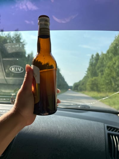 A person's hand holds a brown glass bottle of beer inside a vehicle. The bottle has a label with Russian text and a depiction of a hop cone. The hand is positioned in the foreground, obscuring part of the bottle's label. The vehicle's dashboard is visible below the hand. Through the windshield, a paved road stretches into the distance, lined by green trees on both sides. The sky is clear and blue with some wispy clouds. The overall impression is that the vehicle is in motion, traveling along a rural road under daylight. The location, Беркуты, Russia, is mentioned in the prompt, suggesting the setting.