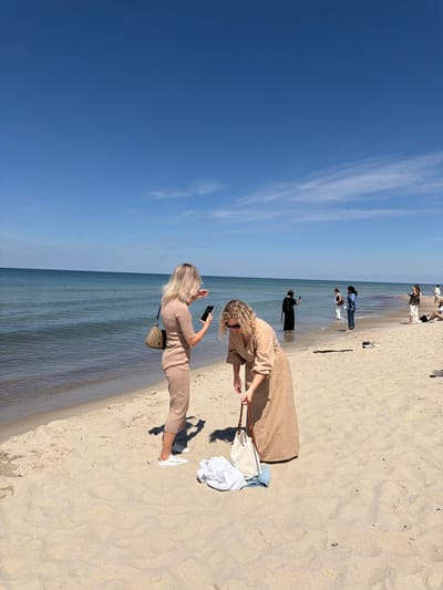 A group of people are on a sandy beach with a calm blue ocean in the background under a clear blue sky. In the foreground, a woman wearing a light brown dress and white sneakers is bending down, holding a beige handbag. To her left, another woman in a light brown ribbed dress and white sneakers is standing and looking at her phone. In the middle ground, several other individuals are visible near the water's edge. One woman in a black dress is taking a photo with her phone, while another in a denim jacket and white pants stands in the water. Further back, a woman in a green shirt and white skirt is also visible. The scene appears to be a leisurely day at the beach in Rybachy, Russia.