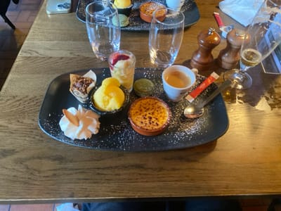 The image displays a table setting featuring a dessert course and beverages in an indoor establishment located in La Wantzenau, France. A central black rectangular plate, dusted with powdered sugar, holds an assortment of individual desserts: a crème brûlée in a terracotta ramekin, a dollop of white whipped cream garnished with a light-colored biscuit wafer, two scoops of yellow sorbet or ice cream in a small black bowl, a mini muffin in a patterned paper liner, a clear shot glass containing layered fruit with a red topping, and a single green macaron. Also positioned on this plate are a white espresso cup containing coffee, a red and white sugar packet, and a silver spoon.
Adjacent to the main plate, on a visibly grained wooden table surface, are two wooden grinders (presumed salt and pepper shakers), a clear stemmed glass holding a small quantity of light amber liquid (likely beer), and two clear water glasses (one full, one with residual liquid). An iPhone with a silver back and two camera lenses is visible in the upper left background. Another black dessert plate containing a crème brûlée, a yellow macaron, and an empty clear glass is present in the background. A crumpled white paper item is on the table to the right. No individuals are depicted, but their presence is inferred by the dining arrangement.