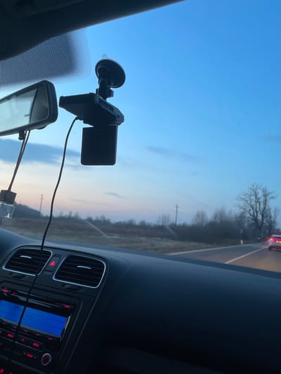 The image depicts a forward-facing view from inside a vehicle, likely in motion, within the city of Переславское, Russia, during dusk or dawn. The upper portion of the frame shows a gradient sky transitioning from light blue overhead to faint pink and orange hues near the horizon.
Within the vehicle's interior, the dark dashboard occupies the lower half of the image, featuring air conditioning vents, a red hazard light button, and a car stereo unit with a blue illuminated "AUX" display and red-lit buttons. A rearview mirror is visible on the left side of the windshield. A black dashcam, attached to the windshield via a suction cup mount, hangs prominently in the upper-center, with its screen oriented downwards. A black power cable extends from the dashcam, and another cable with a clear USB power adapter hangs near the rearview mirror.
Outside the vehicle, a two-lane asphalt road extends into the distance, with a solid white line delineating the right shoulder. A white or light-colored car with illuminated red taillights is visible ahead on the road. The immediate roadside consists of dry, light-colored vegetation and bare trees, indicative of a colder season. Utility poles and additional sparse trees line the horizon, suggesting a rural or semi-rural environment. No individuals are visible in the scene.