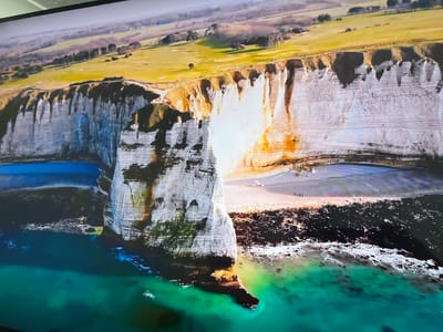 The image shows a coastal landscape, specifically the cliffs of Étretat in Normandy, France, not Basingstoke and Deane, United Kingdom. The prominent feature is a large, natural chalk arch formation extending into the sea, partially covered in vegetation. The water near the cliffs is turquoise in color. Above the cliffs, there is a grassy plateau with a golf course. A small beach is visible at the base of the cliffs with a few people on it. The top of the cliffs appear to have people walking along them.