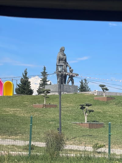 A large, monumental statue dominates a public park in Marneuli, Georgia, under a clear blue sky on a bright, sunny day. The statue depicts a prominent female figure, seemingly leading one or two smaller figures, possibly children, on a raised plinth. The figures are rendered in a realistic, grey stone or metal.
The park features extensive green lawns, neatly maintained with scattered trees; some are distinctly manicured ornamental trees, while others are larger, dark green evergreen trees. A wire mesh fence separates parts of the park, and visible in the background are distant, snow-capped mountains, suggesting the region's mountainous geography.
Overhead, power lines crisscross the sky, adorned with small, triangular decorative flags or bunting, adding a festive touch. In the lower left foreground, parts of a colorful yellow and pink playground structure are visible, indicating the park's recreational use. The scene is devoid of people, presenting a tranquil view of the public space. No discernible text is visible on the monument or its surroundings.