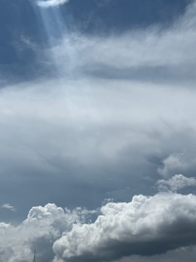 A cloudy sky is depicted in the image, with rays of light breaking through the clouds in the upper left portion. The clouds vary in density and color, ranging from bright white to dark gray, suggesting a dynamic weather system. In the lower portion of the image, a dark, silhouetted object, possibly a mast or antenna, is visible against the bright clouds. The overall scene is of a cloudy sky, likely in the afternoon, with a hint of a structure protruding into the frame from below. The location is Indiantown, United States of America.