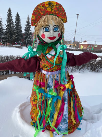 An effigy, consistent with a traditional Maslenitsa doll, stands prominently in a snow-covered outdoor environment in Plast, Russia, under an overcast sky. The doll, facing forward, features a white, spherical head with drawn blue eyes, black eyebrows, red cheeks, and red lips. Its blonde yarn hair is visible beneath a semi-circular headdress adorned with red, yellow, and green floral patterns. A green scarf is tied around its neck.
The effigy wears a long-sleeved, reddish-brown garment with outstretched arms. A light teal ribbon is tied around the left elbow. The torso section of the dress exhibits a reddish-brown and yellow floral pattern. Several small, white rectangular paper tags, possibly containing written text, are attached to the chest, along with a green beaded necklace. A light purple ribbon separates the torso from the skirt. The multi-layered skirt features a reddish-brown and yellow floral pattern, partially covered by a panel of blue and white floral fabric. Numerous lengths of twine hang from the waist, each decorated with multiple colorful satin ribbons tied into bows, including shades of green, blue, yellow, and fuchsia.
The effigy is anchored in a circular impression within the white, undisturbed snow covering the foreground. Behind the doll, low, snow-dusted dark green bushes are visible. In the midground, several tall, dark green coniferous trees stand against the sky. The background reveals a street with a parked white car, various low-rise buildings (some with red roofs), utility poles with overhead wires, and streetlights. Pedestrian crossing signs are also discernible. Distant, indistinct human figures are visible on the street, and colorful flags or banners are present in the far background, suggesting a public area.
