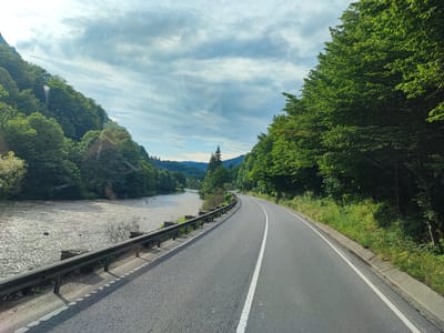 This media file shows a scenic view of a winding asphalt road running alongside a river in a mountainous, forested region. The road has a white line marking its center and edge. The river, which appears to be flowing with some current, is on the left side of the road, bordered by a metal guardrail and some stone embankments. Dense green trees and foliage cover the steep slopes on both sides of the road and river, creating a lush, natural environment. In the distance, rolling hills and mountains are visible under a cloudy sky. The lighting suggests it is daytime, possibly late afternoon, as the sun casts a warm glow on the water's surface. There is no visible text in the image. The scene evokes a sense of peaceful travel and natural beauty, characteristic of a scenic drive through a rural or wilderness area. The location is identified as Andreneasa, Romania.