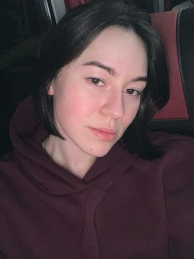 A woman with dark hair and light skin, clad in a maroon hoodie, reclines against what appears to be a dark surface and a red cushion. Her face is tilted slightly towards the camera, revealing soft features and brown eyes. A delicate silver necklace is visible at her collarbone. The lighting is soft, emphasizing the texture of her skin and the cozy material of the hoodie.