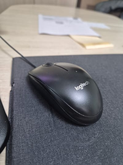 The media file is a close-up, eye-level shot of a black Logitech computer mouse resting on a dark grey mousepad. The mousepad is textured and appears to be on a wooden desk. To the left of the mousepad, a light-colored surface, also likely a desk, is visible. On this surface, there is a white piece of paper with some faint markings and a small, folded yellow sticky note. The scene suggests a typical home or office workspace. The lighting appears to be ambient, and there are no strong shadows, indicating it's likely daytime or a well-lit indoor environment. The focus is sharp on the mouse and mousepad, with the background slightly blurred, drawing attention to the primary object. The overall impression is one of a functional workspace. Visible text on the mouse reads "Logitech". The context provided indicates the location is Vileyka, Belarus. No people are visible in the image.