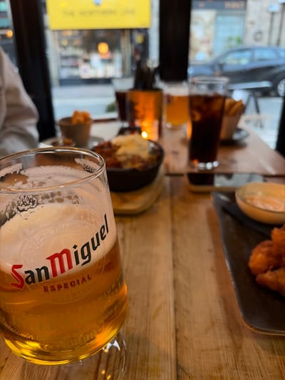 Here's a description of the image:
The photograph is taken from a low angle, at table level, likely inside a restaurant or pub, with a focus on a partially consumed glass of "San Miguel Especial" beer. The beer glass is prominently positioned in the foreground. The beer has a light golden color and a frothy head. The San Miguel logo is clearly visible on the glass.
The table appears to be made of wood, with a warm, brown tone. Beyond the beer glass are other glasses containing liquid; one with a dark color (possibly a cola) and another with an amber liquid (perhaps orange juice or another beer). Further in the background, there are dishes with food; a dark tray with fried food and a bowl.
Through the window, a blurred cityscape is visible. A storefront with a yellow sign is partially visible, and the reflection of a car can be seen in the window. The scene suggests a casual dining or pub atmosphere. The photo captures a moment of enjoying food and drinks, likely during a meal or social gathering. There are no people visible. The setting is most likely The Northern Line in Windermere, United Kingdom, based on the words on the yellow sign.