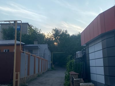 The image depicts an urban scene in Antratsyt, Ukraine. The photograph is taken from a street-level perspective, looking down a narrow alleyway lined with buildings and a fence. The sky above is partially cloudy, with thin, wispy clouds and some sunlight filtering through the trees in the background, suggesting it is either early morning or late afternoon.

On the right side of the alley, a modern building with a red-toned facade and a gray and white tiled exterior wall is visible. An air conditioning unit is mounted on the exterior. On the left side of the alley, there are several structures. Closest to the viewer is a dark brown fence, followed by a fence with orange and gray panels supported by vertical posts. Beyond this fence, a low-rise building with an orange facade and a dark roof is visible, with a window and part of a white door. Further down the alley, a light-colored building with a dark roof and a balcony is seen, with trees in front of it.

In the background, the alleyway curves and leads towards a darker area where a car is parked. Trees densely populate the area behind the buildings, with the setting or rising sun peeking through the leaves, creating a dappled light effect. Yellow pipes, possibly for utilities, are visible running along the fence and across the alley. The ground surface of the alley appears to be asphalt or a similar paved material. There are no people visible in the image.