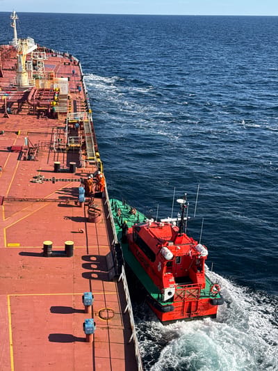 An elevated, extreme wide shot captures a pilot boat, painted bright orange and green, maneuvering alongside a large red tanker ship. The pilot boat is leaving a wake in the deep blue ocean. The tanker's deck is red and marked with yellow lines, featuring numerous pipes and equipment. Several individuals in orange worksuits are visible on the tanker's deck, near the edge where the pilot boat is positioned. The sky is a clear, pale blue. The scene appears to be in open water, with no discernible urban or land features visible that would indicate a specific city in Denmark.