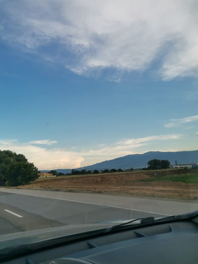 The image captures a view from a vehicle traveling along a paved road. The road has a white line marking the lane. In the foreground, the dashboard and windshield wipers of the vehicle are visible. The landscape to the right of the road features dry, brown earth with some scattered green vegetation and a few scattered trees. In the middle ground, there is a building with an orange facade and a flat roof, surrounded by vegetation. Further in the background, a range of dark blue mountains stretches across the horizon. The sky is a clear blue with scattered white clouds, some of which are illuminated by sunlight, creating a bright, somewhat hazy effect. The overall scene suggests an outdoor environment, possibly a rural or semi-rural area, with evidence of construction or land preparation in the mid-ground. The location is identified as Krivina, Bulgaria.