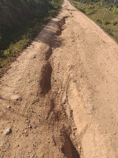 A dirt road angles sharply to the left, flanked by green vegetation. The road surface is uneven, with ruts and shadows indicating erosion. A small, straight stick lies on the road, casting a distinct shadow. The terrain appears rural, likely a pathway through a natural area. The lighting suggests it's either early morning or late afternoon.