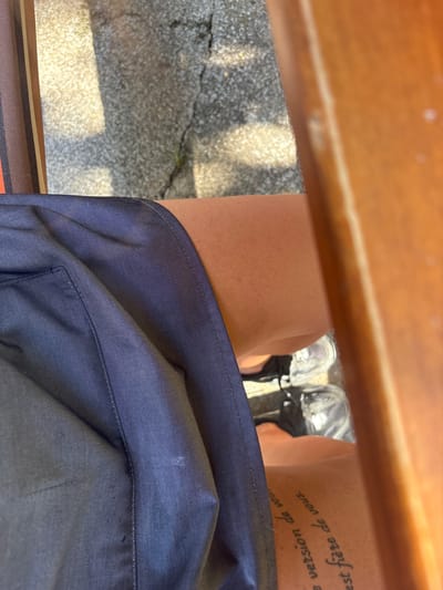 The image, captured from a first-person, downward-looking perspective, depicts the lower body of an individual seated outdoors. The subject wears dark blue shorts or a skirt made of a finely ribbed fabric, covering the upper thighs. The exposed skin of the thighs is of a medium tone, with a cursive tattoo visible on the right thigh, displaying partial French text: "e version de vou" and "est fière de vous." On the feet, light grey and black athletic shoes with white soles are visible, resting on a textured, light grey concrete or asphalt surface. This ground shows multiple cracks and sparse patches of green vegetation. Strong, direct sunlight illuminates the scene, casting distinct shadows on the pavement. To the right, a smooth, light brown wooden surface, possibly part of a chair arm or bench, is present. On the left, a striped pattern in shades of brown, orange, and cream, consistent with upholstered furniture, is partially visible. There is no visual information within the image to specify the location as Hrishteni, Bulgaria, or any other particular city or country.