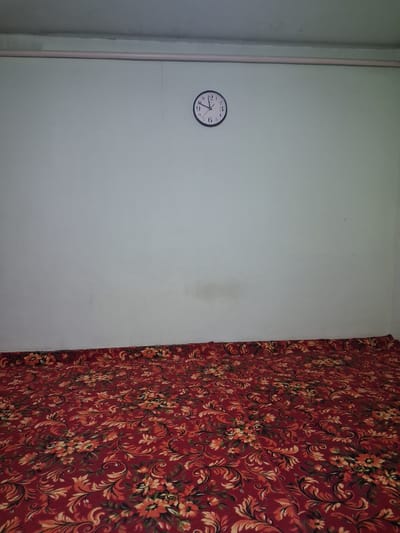 The image depicts an interior scene, likely a room, with a light-colored wall and a patterned red carpet covering the floor. A round clock with black numerals and hands is mounted on the wall. The clock appears to show a time between 10:00 and 10:05. A thin, pinkish pipe runs horizontally across the upper part of the wall. There are no people or visible actions in the image. The environment is sparsely furnished, with the focus being on the wall, clock, and carpet. The overall impression is one of simplicity and a lack of activity. This scene is located in Kahramon, Uzbekistan.