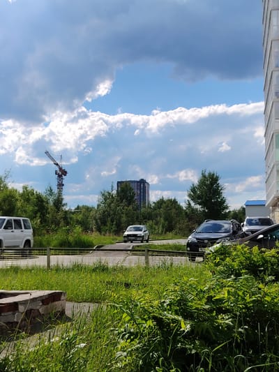 In Pyt'-Yakh, Russia, a street-level view captures a cloudy sky with patches of blue. In the distance, a red construction crane stands tall against the sky, accompanied by a dark, multi-story building. Closer to the foreground, several cars are parked on the side of a road, with lush green grass and trees bordering the scene. A white SUV is parked on the left, and a dark SUV is parked further down on the right. A portion of a modern apartment building is visible on the far right edge of the image. In the immediate foreground, a low brick structure and dense greenery are present.