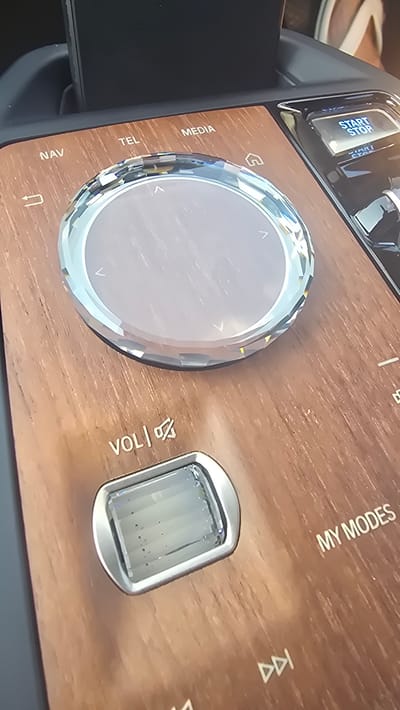 The image shows a close-up of a car's center console, specifically the dashboard area. The console features a wooden-grain finish. Prominently displayed is a large, crystal-like rotary dial with directional arrows around its edge, likely for navigation or media control. To the left of the dial, there are illuminated buttons labeled "NAV," "TEL," and "MEDIA." To the right of the dial, a blue illuminated button reads "START/STOP." Below the rotary dial, a smaller button with a crystal-like insert is labeled "VOL/MUTE." Further to the right, the text "MY MODES" is visible, along with media playback icons. The background is blurred but suggests the interior of a vehicle. The scene is static and shows no people or immediate actions. The context of Parekklisia Community, Cyprus, is not directly inferable from the visual elements of the image itself but is provided as external information.