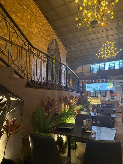 The image depicts the interior of a restaurant or cafe, likely in Ban Na Chom Thian, Thailand, given the location context. The setting is dimly lit, suggesting it is evening or the interior lighting is low. The scene is dominated by a grand staircase with intricate metal railings and a brick wall, leading up to a large, arched window with stained glass. Above the staircase, two ornate chandeliers with many yellow lights hang from a metal-gridded ceiling.
Several tables with chairs are visible in the background, some occupied. The tables are dark, and on one table, there are glasses and a cup. Green plants with large leaves and some with red foliage are strategically placed around the seating area, adding a touch of natural decor.
In the middle ground, a digital screen displays images and text. One screen shows food, another displays a drink, and a third prominently reads "NOW SOFT MATCHA". Small rainbow-colored flags are also visible, possibly indicating a Pride Month celebration or general inclusivity. The overall atmosphere appears relaxed and inviting, with a focus on ambiance and dining experience. The presence of both indoor plants and artificial lighting creates a cozy, yet sophisticated, environment.