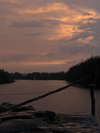 This image depicts a serene sunset over a body of water, likely a river or a lake, in Rhieng Krueng, Indonesia. The sky is painted with hues of orange, pink, and purple as the sun begins to set, casting a warm glow on the clouds. The water reflects the colors of the sky, creating a tranquil atmosphere.
In the foreground, a pile of large, dark rocks occupies the lower portion of the image, leading the viewer's eye towards the water. A rustic wooden structure, possibly a pier or a part of a fishing platform, extends from the right side of the frame, with a rope tied around a wooden post.
The middle ground features the calm water, with faint ripples suggesting a gentle breeze. On the far bank, a dense line of tropical trees and vegetation forms a dark silhouette against the vibrant sky. A small, elevated structure, possibly a lookout post or a shelter, can be seen on the left bank, partially obscured by foliage. A few small birds can be seen flying in the distance, adding to the natural ambiance of the scene. The overall mood is peaceful and reflective, capturing the beauty of nature at dusk.