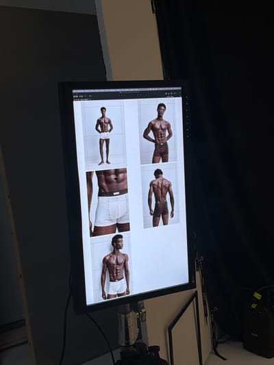 The image depicts a professional photography setup, likely in a studio. A large monitor displays a grid layout with multiple images of a male model posing in underwear. The model is featured in various poses, including front, side, and back views, with close-ups of his torso and lower body. The monitor is mounted on a stand, suggesting it's used for reviewing and editing photographs. In the background, studio lighting equipment and dark fabric backdrops are visible, indicating a controlled shooting environment. The overall scene conveys a professional and organized atmosphere associated with a photoshoot.