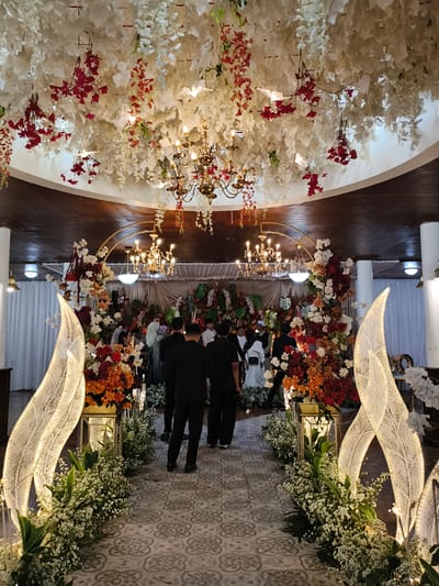 This detailed image, captured in Sindanglaya, Indonesia, depicts an indoor formal event space adorned with extensive decorations. The ceiling features a dense canopy of artificial white and red flowers and leaves, from which several elaborate gold chandeliers with illuminated bulbs are suspended. A patterned floor with grey and white geometric designs forms a central aisle leading towards a heavily decorated background. Flanking this path are large, illuminated, white, leaf-like structures with delicate lace-like patterns, accompanied by low floral arrangements of small white flowers and green foliage. Further along the aisle, larger floral displays in red, orange, and white are placed on gold stands, creating an archway leading to the main area. Several individuals, predominantly men in dark suits, are seen walking along the path towards the background. In the distance, the main area is densely decorated with large floral arrangements and a prominent golden arched structure, with more people gathered. White curtains and pillars line the perimeter of the room, and the overall lighting is dim, accentuated by the warm glow of the chandeliers and decorative lights.