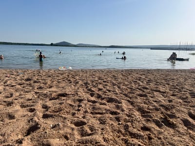The image depicts a sunny day at a beach on a lake, with a clear blue sky and calm water. The foreground is dominated by coarse sand, showing footprints and a few small pebbles. In the mid-ground, several people are enjoying recreational activities in the lake. Some are swimming or wading, while others are floating on inflatable rings and boards. A young person in the water is holding a round inflatable object. Further out on the lake, more individuals are visible, some with floats. In the distance, a tree-lined shore stretches across the horizon, with gentle hills rising behind it. To the right, a marina with numerous sailboats is docked, with their masts creating a distinct silhouette against the sky. The overall atmosphere is one of leisurely summer enjoyment. The time of day appears to be late afternoon or early evening, given the warm, soft lighting and long shadows cast on the sand. The location is Nonsard-Lamarche, France. There is no visible text in the image.