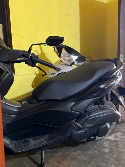 A black Yamaha NMAX scooter is parked in front of a yellow wall in a garage or covered area. The scooter is partially obscured by another motorcycle with a white front fairing. The floor is concrete and appears worn. A dark wooden door is visible on the right side of the image. The scene is consistent with a residential setting in Tanjung Pura, Indonesia, where motorcycles are a common mode of transportation.
