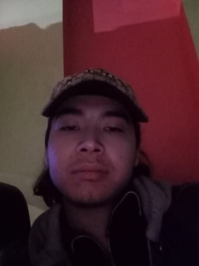 A close-up, head-and-shoulders photograph depicts a single adult male within an indoor setting in Takhtakuprik, Uzbekistan. The subject, of apparent East Asian or Central Asian ethnicity, occupies the central frame, looking directly forward with a neutral expression. He wears a multi-patterned baseball cap, a dark outer jacket, and a grey hooded sweatshirt visible beneath the jacket. Sparse facial hair is present on his chin and upper lip. The background features a wall divided into two distinct color sections: light tan on the left and solid red on the right. The scene is dimly lit, with a subtle blue light source illuminating the left side of the subject's face. No other individuals or significant objects are discernible.