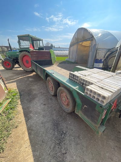 The image depicts an outdoor agricultural or semi-rural scene in Krško, Slovenia, under clear daylight conditions. In the left-center, a green and red agricultural tractor, likely a Fendt, is positioned, with its rear left wheel prominently visible and painted red. The tractor is hitched to a two-axle, green flatbed utility trailer. The trailer's right side is closer to the viewer.
Stacked on the rear half of the trailer's flatbed are numerous rectangular white or light gray concrete blocks, each featuring multiple circular holes. There are approximately five rows of these blocks visible, stacked two or three high. A small dark object, possibly a smartphone, rests on the forward section of the trailer bed.
To the right of the tractor and trailer, a large, translucent plastic-covered hoop-style greenhouse is visible, with its entry flap partially open, revealing a yellow interior. Adjacent to the greenhouse, coiled black hoses and a blue and white barrel (labeled "FUCHS") are present.
The ground is a mix of dirt and gravel, showing tire tracks. In the background, beyond the main greenhouse, more similar greenhouses are visible, alongside what appears to be a field or cultivated land. A few bare trees and distant structures are also present under a bright blue sky with sparse white clouds. No individuals are visible in the scene.