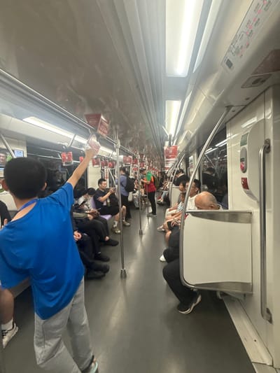 The image depicts the interior of a subway car in Changning District, China, during daylight hours. The car is moderately crowded with passengers seated and standing. A young boy in a blue t-shirt and grey pants is in the foreground, facing away from the camera, with his right arm raised and holding onto a handrail. Several other passengers are visible, including individuals looking at their phones, reading, or resting. The environment is functional and clean, with overhead lights illuminating the carriage and advertisements displayed above the windows. The subway car has a modern design with white interior walls and grey flooring. The overall scene suggests a typical daily commute on public transportation.