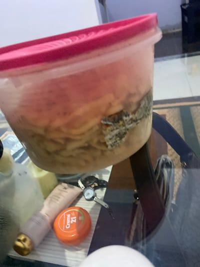 The image shows a glass-top table with various items. In the foreground, a pink-lidded plastic container holds what appears to be food, possibly pasta, with some discolored patches on the side. To the left and slightly behind the container are bottles and tubes of cosmetic products, including a pink tube and an orange round container labeled "Creme 21". A watch with a white face and a metal band is also visible on the table. The background is blurred but suggests an indoor setting with a rug and furniture. The scene is located in Walennae, Indonesia.