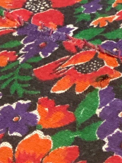 The image is a close-up, high-angle photograph of a floral patterned fabric with a dark background. The pattern consists of large, stylized flowers in red, orange, and purple, interspersed with green leaves. There is a small, dark insect, possibly a fly, perched on one of the orange flower petals in the center-right of the image. The fabric appears to be textured, and the colors are vibrant. The photograph was taken in Arbagish, Uzbekistan.