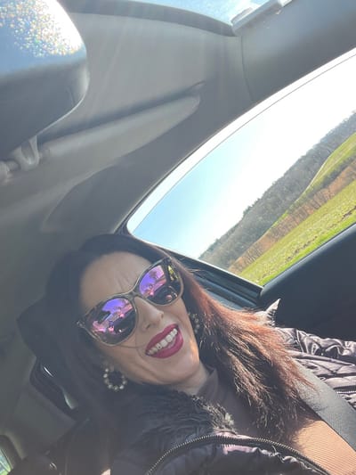 An adult female is depicted inside a vehicle, occupying a front seat, in a location consistent with a road near Olingen, Luxembourg. She is seen from the chest up, smiling directly at the camera. She wears large, reflective, purplish sunglasses, obscuring her eyes. Her mouth is open, revealing white teeth and a small stud on an upper incisor, accentuated by dark red lipstick. She has long, dark reddish-brown hair. Her left ear displays a large, circular, possibly pearl-studded earring. She is dressed in a dark, quilted jacket with a fur-like collar, over a brown ribbed top, secured by a black seatbelt across her chest. The vehicle's interior features a light-colored headliner and a rearview mirror visible in the upper left, with bright sunlight reflecting off it. Through the passenger side window on the right, a clear blue sky is visible above rolling green fields and a distant treeline composed of what appear to be deciduous trees without leaves. The reflections in her sunglasses show elements of the car's interior and the external landscape.