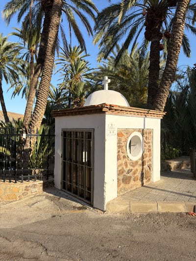 The image shows a small, white structure with a domed roof topped by a cross, located in the Sierra Alhamilla region of Spain. The structure is situated outdoors, surrounded by palm trees and vegetation. The front of the structure features a section of stonework and a small window with a metal grate. A sign is affixed to the wall near the window, bearing text in Spanish. A black metal fence with stone supports borders the area to the left. The ground in front of the structure is paved with asphalt and some of the pavement is cracked. The lighting suggests it is daytime, with sunlight casting shadows.
