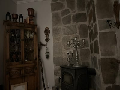 This image depicts a dimly lit interior space, likely a room within a dwelling in Gränsta, Sweden. The scene is static, featuring no individuals.
To the left, a tall, antique-style wooden display cabinet stands against a light grey wall. The upper section of the cabinet has two glass-fronted doors with diamond-patterned mullions, revealing various objects including a white bust and bottles on shelves. The lower section consists of two solid wooden doors with carved details. On top of the cabinet, from left to right, are two black decorative lanterns, a smaller wooden drum, several stacked rectangular items (possibly books), and a larger wooden drum. A light-colored, empty picture frame leans against the cabinet's base on the floor.
The light grey wall meets a corner where a prominent feature wall constructed of irregularly shaped, mortared stone blocks dominates the right side of the frame. On the grey wall, an ornate, dark, carved wooden wall plaque is mounted. Below it, a tall staff-like object with a curved, horn-like top leans against the wall. A small, dark hanging lantern with metal supports is suspended from the wall below the plaque.
Positioned in the corner, in front of the stone wall, is a dark, cast-iron wood-burning stove with a visible front door. On top of the stove, a silver-colored candelabra with five candle holders stands centrally, holding three white candles. To its left, a smaller dark metal candle holder with two white candles and two circular loops is present. Further up on the stone wall, a dark metal sconce with a single candle holder is mounted, and a small dark decorative element hangs lower on the same wall.