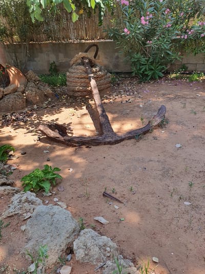 An old, rusted anchor is displayed on sandy ground in Malia Municipal Unit, Greece. The anchor is positioned horizontally, with its flukes pointing to the right and its shank extending towards the left, where it is secured by a thick, coiled rope. Behind the anchor, a concrete wall runs across the scene, topped with a woven bamboo fence. To the left of the anchor, a large terracotta pot with some plants and pink flowers is visible, along with several large rocks. To the right, a dense green bush with pink flowers provides a backdrop. The ground is covered with sand, scattered leaves, and small green shoots. Sunlight casts shadows across the scene.