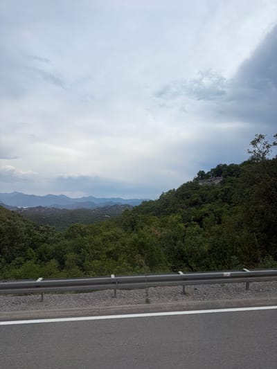 This media file depicts a scenic mountain landscape in Gospoština, Montenegro, viewed from the side of a road. The image is oriented vertically. On the right side, a paved road with a white line marking is visible, bordered by a metal guardrail and a gravel shoulder. To the left of the road, a steep, densely forested mountainside rises, covered in lush green trees.
In the distance, a series of layered mountain ranges recedes into the haze, creating a sense of depth and vastness. The sky is filled with dramatic clouds, with patches of brighter sky peeking through. The overall atmosphere is serene and natural. There are no people or specific activities depicted. The time of day appears to be daytime, possibly overcast or late afternoon, given the diffused light. The location is identifiable as Montenegro due to the provided context. There is no visible text in the image.