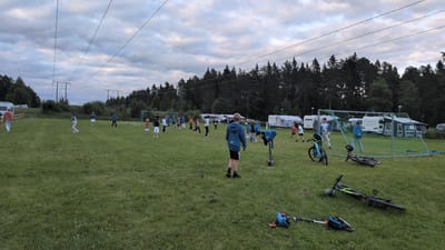 The image depicts an outdoor scene in Östersund, Sweden, likely during late afternoon or early evening due to the dim lighting and cloudy sky. A group of people, predominantly children and teenagers, are gathered on a grassy field. Some individuals appear to be engaged in a soccer game, with a soccer goal net set up towards the right side of the frame. Several white camping trailers and awnings are visible in the background, suggesting the location might be a campground or an event area. Power lines are strung across the sky, supported by tall poles. In the foreground, two bicycles and a scooter are present on the grass, one bicycle lying on its side. The environment is characterized by a large, open grassy area surrounded by a dense line of trees in the distance.