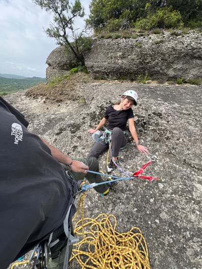 This image captures a rock climbing scene from a first-person perspective, looking upwards at a climber on a rocky cliff. The overall image is rotated 90 degrees counter-clockwise.
The primary subject is a woman, wearing a white climbing helmet, a black short-sleeved top, and dark grey climbing pants. She has a harness on with various climbing equipment attached, and is wearing climbing shoes. She is seen resting on the rough, grey rock face, with her left hand placed on the rock. She is smiling and looking towards the camera. Two red quickdraws are visible near her, attached to the rock and the rope system.
In the foreground, partially visible, is the arm and torso of another person, presumably the belayer and photographer. This person is wearing a dark grey t-shirt with the upside-down logo "THE NORTH FACE" printed in white. Their left hand is holding a blue and white patterned climbing rope. A yellow climbing rope, coiled neatly, rests on the rock surface to the right of the belayer's position. The belayer's harness and other gear are also partially visible.
The setting is a rugged outdoor environment, likely a mountainous or cliffside area. Beyond the rock face, green trees and vegetation can be seen, with a hazy, overcast sky and distant mountains or hills in the background. The rock itself is textured, with lighter and darker patches, and some small green plants growing on it.
Notable details include the text "HAPPY CLIMBER PETZL" visible on the woman's helmet. The scene depicts an active rock climbing session, likely in Kastraki, Greece, given the location context. The overcast sky suggests daytime.