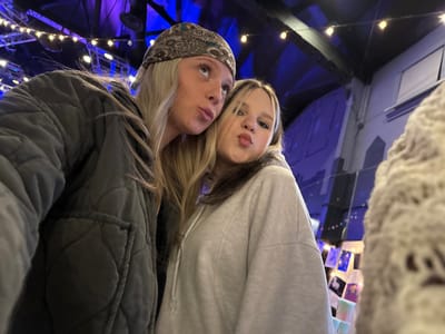 Two fair-skinned women pose closely together in an indoor space lit with blue and white lights. The woman on the left wears a light gray hooded sweatshirt. The woman on the right sports a quilted olive green jacket and a patterned headband. The room's architecture suggests a theater or performance venue with visible support structures, string lights, and photo displays. A blurry, textured object appears on the far left edge of the frame.