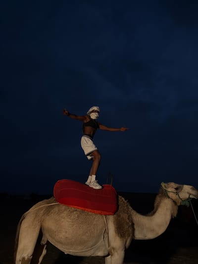 A person wearing a white head covering and face mask stands with arms outstretched on the back of a camel. The person is dressed in a black sports bra and white shorts. The camel is equipped with a red saddle. The scene is set at dusk in a dark, open area, possibly the desert, with a dark blue sky above. In the background, there are faint outlines of structures, suggesting a settlement. The overall impression is of a performance or acrobatic display.
