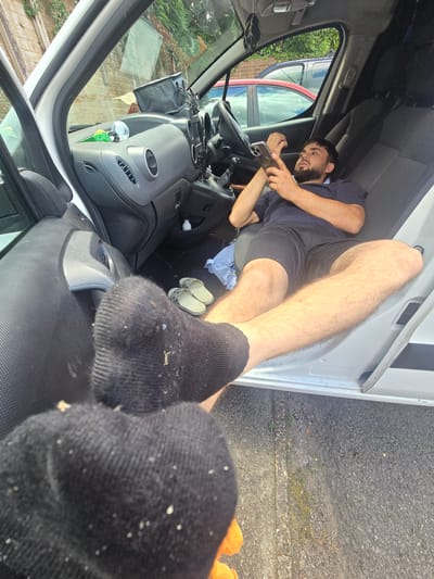 In Woking, United Kingdom, a man is lying down inside a white van, holding a smartphone. He is wearing a dark blue t-shirt and black shorts. His legs are extended and bare. In the foreground, a black sock is visible, with some debris on it. Outside the van, a paved surface with some vegetation is visible. The van's interior shows seats, dashboard, and a steering wheel. There are also a pair of sandals on the floor of the van. The scene appears to be set in an outdoor environment, possibly a street or parking area, given the presence of the paved ground and vegetation.