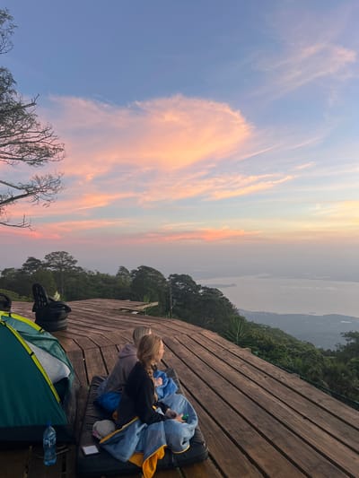 The image captures a serene outdoor scene at El Cacao, El Salvador, likely during sunrise or sunset given the soft, pastel hues of the sky. The sky is a canvas of gentle blues and oranges, with wispy clouds stretching across. In the foreground, a wooden deck serves as the setting for a campsite. A green tent is partially visible, along with a blue plastic water bottle. Two individuals are present on the deck, nestled amongst sleeping bags and mats. One person, with blonde hair, is looking out at the landscape, while another individual is partially visible beside them. They appear to be resting or enjoying the view, conveying a sense of peace and tranquility. Beyond the deck, lush green trees and hills dominate the landscape, with a body of water and distant landmasses visible on the horizon, shrouded in a soft haze. The overall mood is one of calm and appreciation for nature's beauty.
